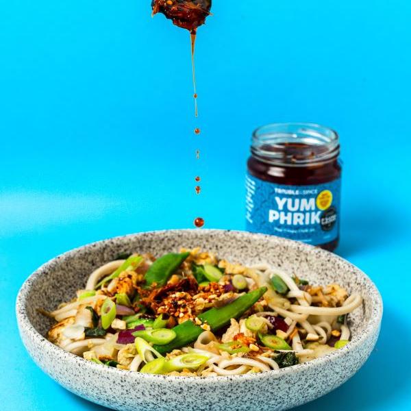 Bowl of noodles with vegetables being drizzled with Yum Phrik crispy chilli oil - chili olie, with the jar in the background against a bright blue backdrop