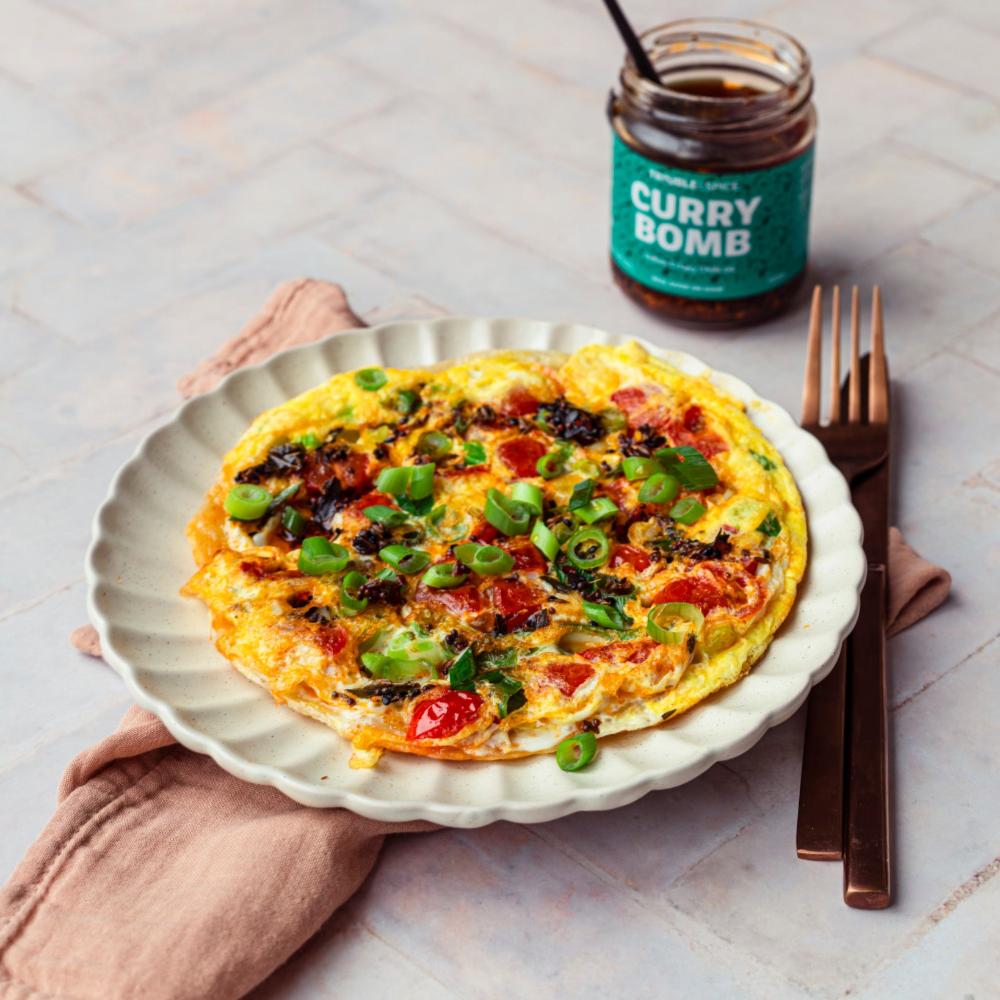 Paratha omelette topped with chopped green onions, tomatoes, and a drizzle of Curry Bomb Indian crispy chilli oil – chili olie, served on a scalloped plate with a jar of Trouble & Spice Curry Bomb in the background.