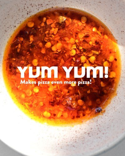A close-up of a bowl filled with vibrant chilli oil (chili olie) used as a spicy pizza sauce. Text overlay reads “YUM YUM! Makes pizza even more pizza!”.