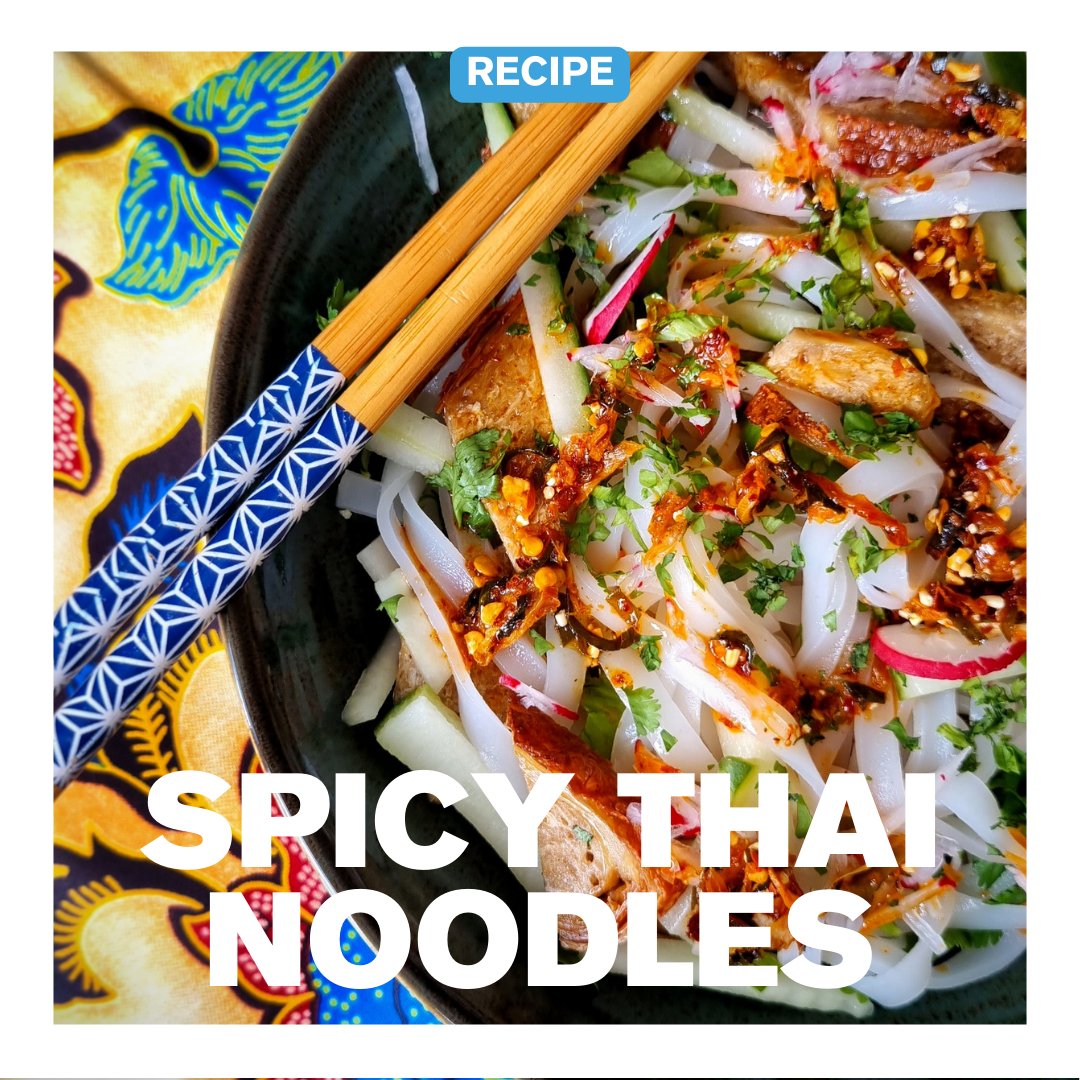 Spicy Thai noodles topped with crispy tofu, herbs, and drizzles of crispy chilli oil – chili olie, served in a dark bowl with chopsticks on a vibrant batik cloth.
