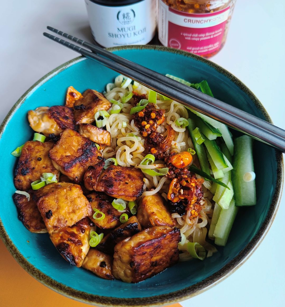 Bowl of noodles topped with crispy tofu, cucumber, and Trouble & Spice Crunchy Spice chilli crisp, served with Mugi Shoyu Koji.