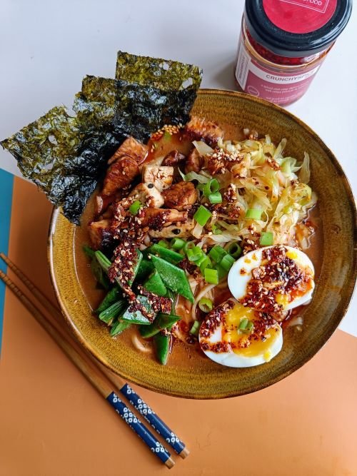 Bowl of ramen topped with soft-boiled egg, green vegetables, shredded chicken, seaweed, and Trouble & Spice Crunchy Spice chilli crisp.