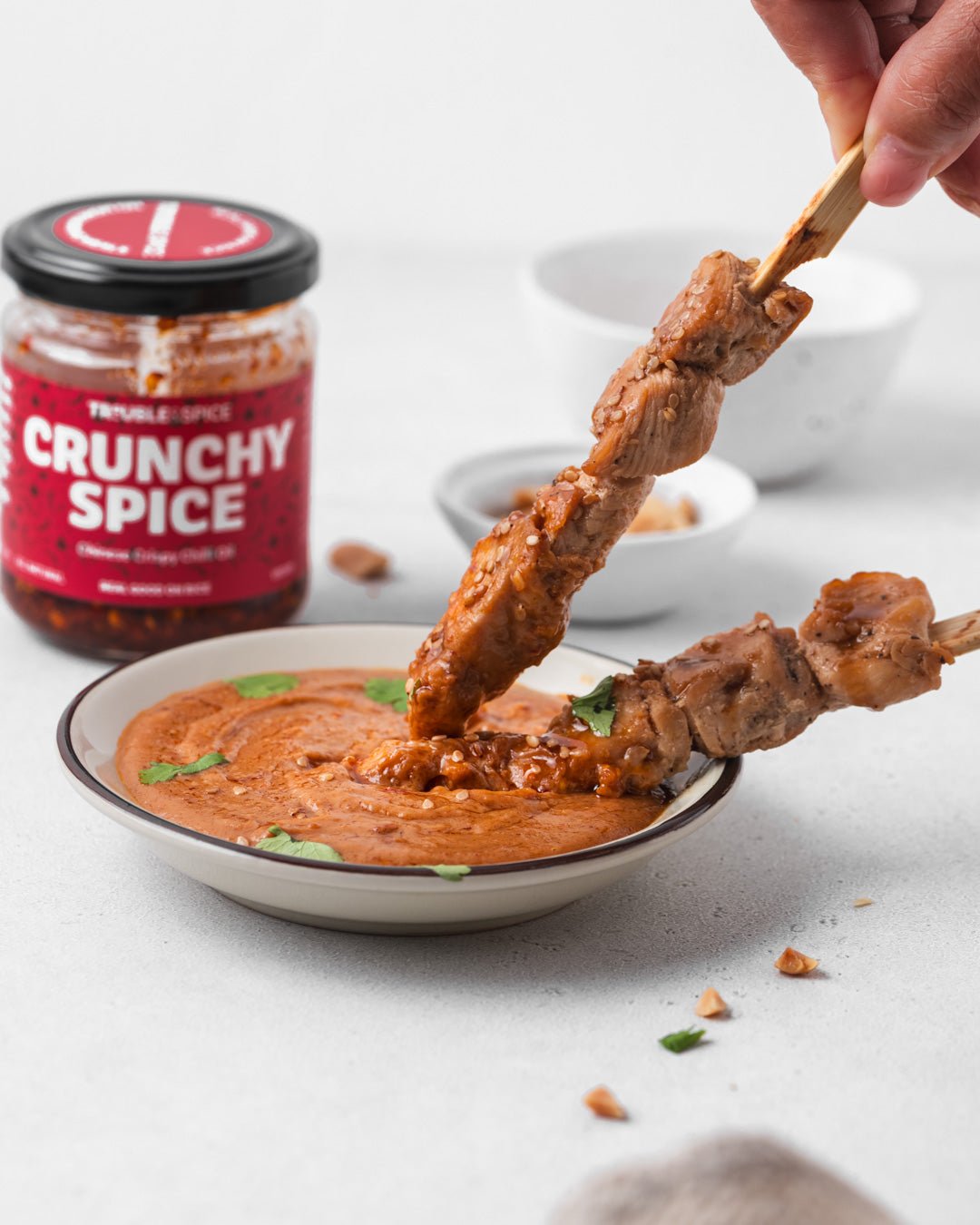 Chicken satay skewers being dipped into creamy peanut sauce, served with Trouble and Spice Crunchy Spice crispy chilli oil - chili olie in the background.