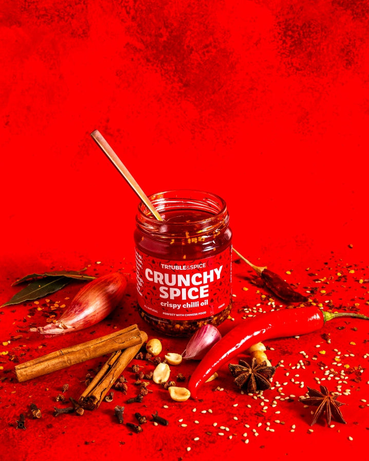 Jar of crunchy spice crispy chilli oil with cinnamon sticks and red peppers on a red background