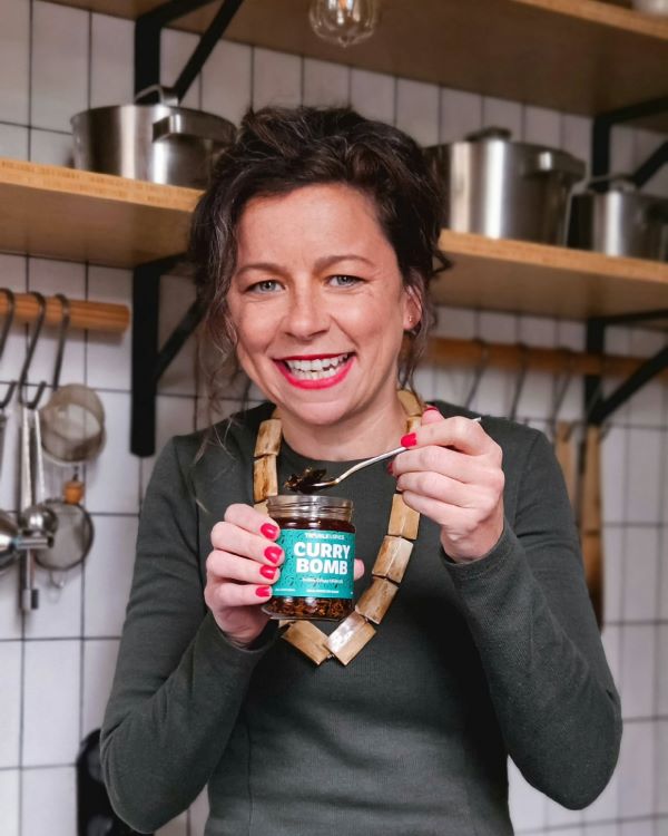Nicole smiling in a kitchen while holding a jar of Curry Bomb crispy chilli oil - chili olie and scooping some with a spoon.
