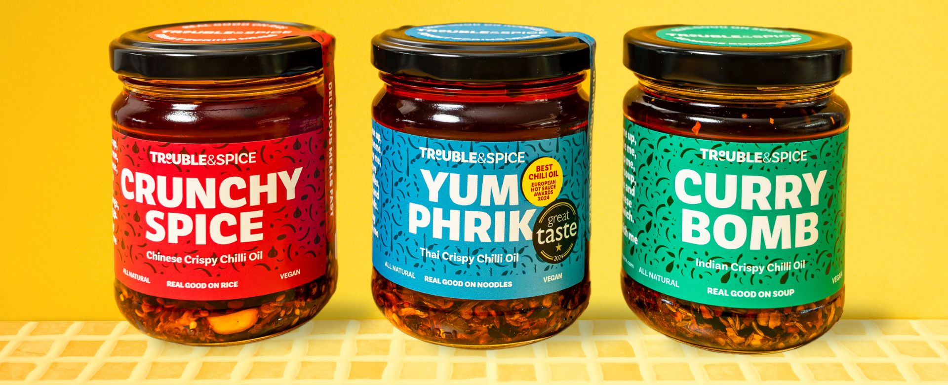 Three jars of Trouble & Spice crispy chilli oils chili olie  displayed side by side against a yellow background.