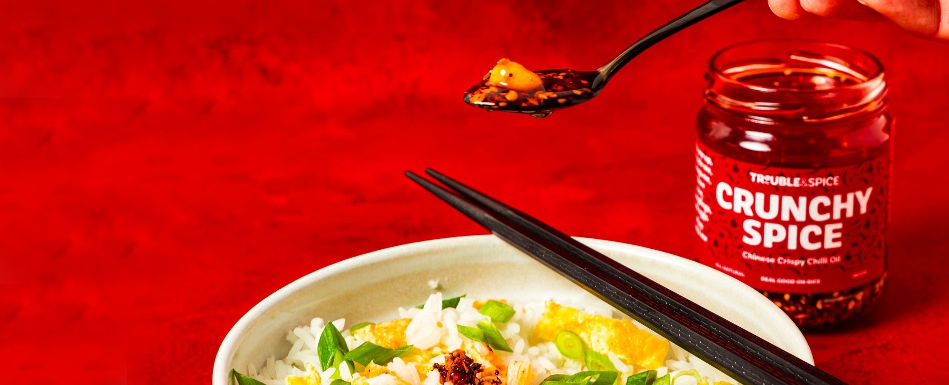 A hand holds a spoonful of Trouble & Spice Crunchy Spice crispy chilli oil - chili olie  above a bowl of rice topped with vegetables and sauce. A jar of the Chinese-style chilli oil is visible in the background against a bold red backdrop.