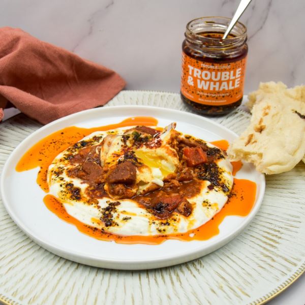 Plate of cilbir shakshuka topped with a poached egg, drizzled with Trouble & Spice Trouble & Whale Turkish crispy chilli oil, served with flatbread and a jar of the chili oil - chili olie in the background.