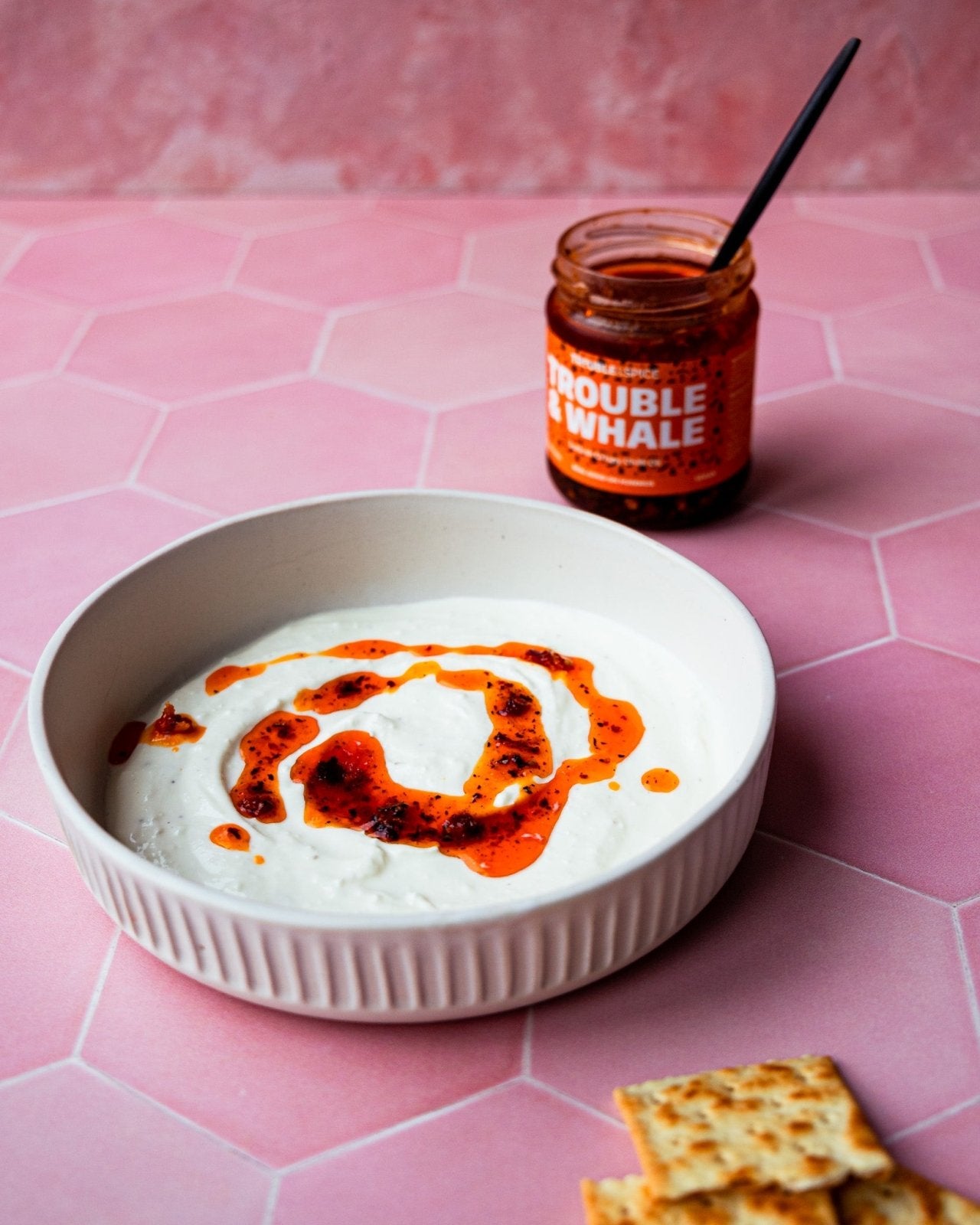 Bowl of creamy labneh topped with a swirl of Trouble & Spice Trouble & Whale Turkish crispy chilli oil - chili olie, with a jar of the chili oil and crackers on the side.