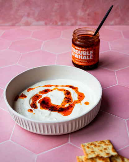 Bowl of creamy labneh topped with a swirl of Trouble & Spice Trouble & Whale Turkish crispy chilli oil - chili olie, with a jar of the chili oil and crackers on the side.